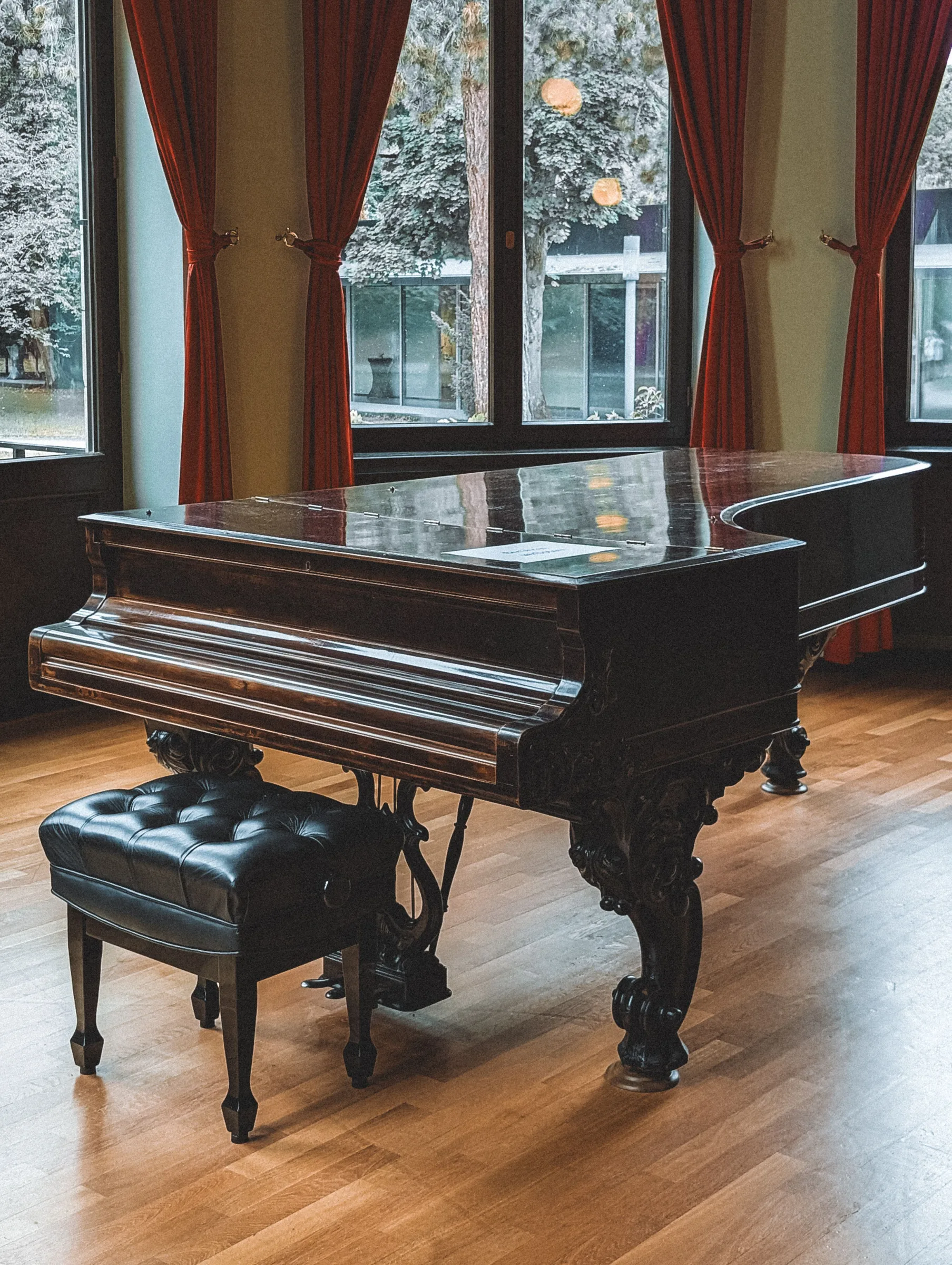 Black grand piano with matching bench in an elegant room with red curtains and large windows.