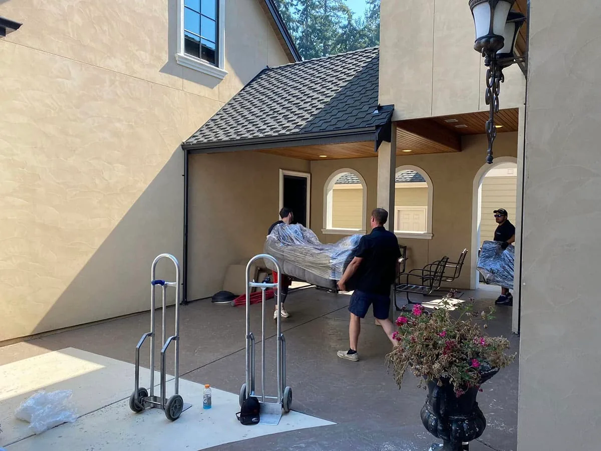 Moving crew unloading wrapped furniture into a residential garage with black roof.