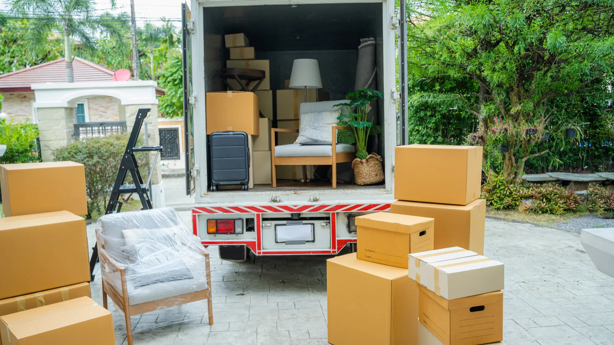 The open back of a moving truck, empty of people, reveals a packed interior: stacked cardboard boxes, a rolled rug, a wooden