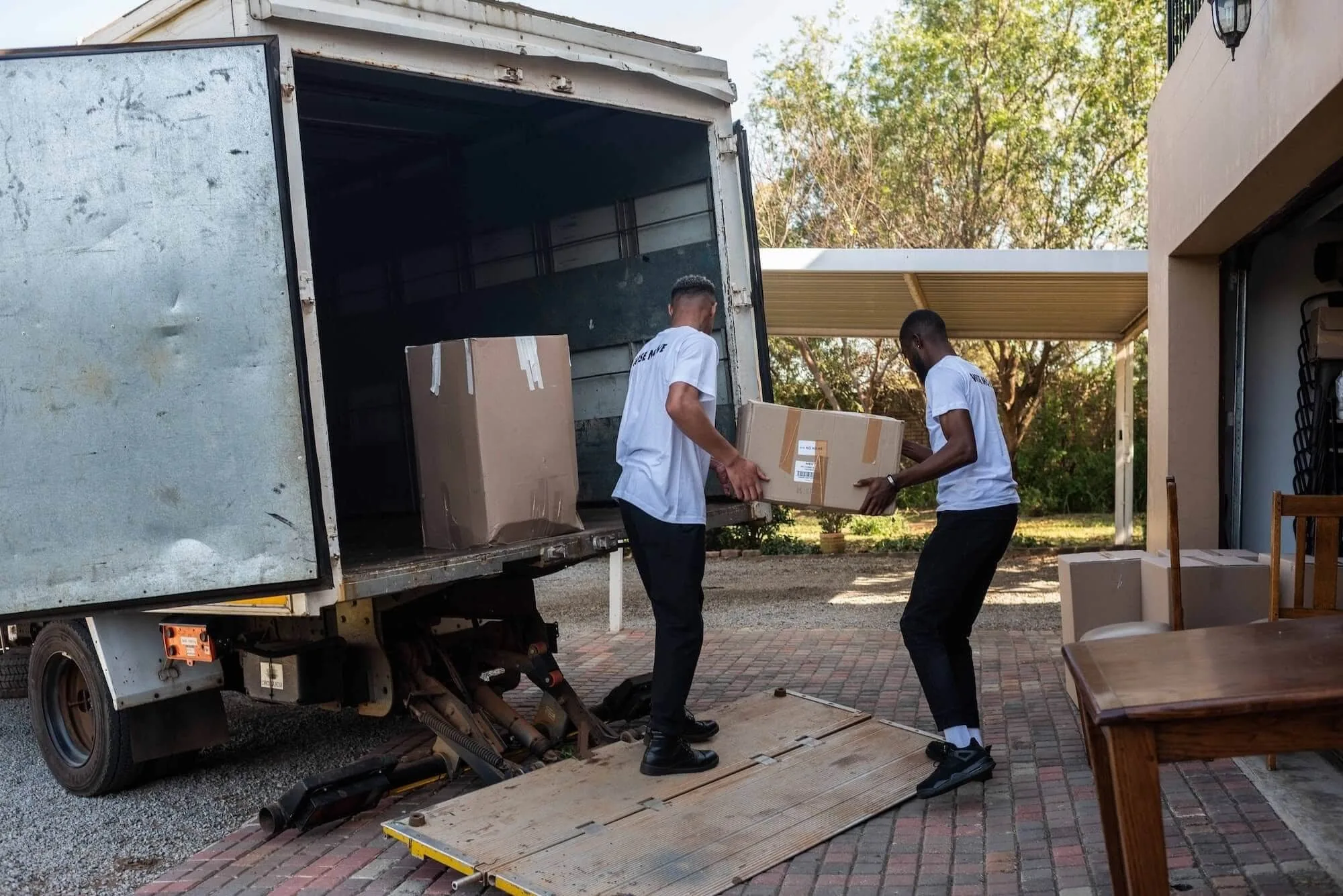 Two movers in white shirts unload cardboard boxes from a moving truck at a residential driveway.